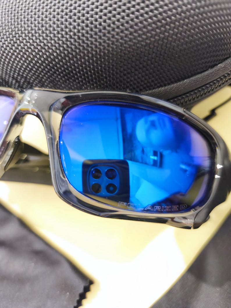 May include: Close-up of polarized sunglasses with blue lenses and a black frame. The lenses reflect a bright blue hue, and the text "POLARIZED" is visible on the lens. The sunglasses are resting on a black case.