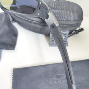 May include: Black sunglasses with a black case and cleaning cloth. The sunglasses have a sleek, modern design with a black frame and tinted lenses. The case is a dark gray, zippered, and the cleaning cloth has the brand name printed on it.
