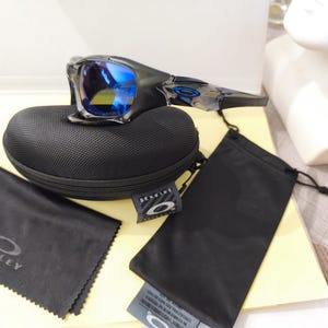 May include: Black Oakley sunglasses with blue mirrored lenses rest on a black case. A black microfiber cleaning cloth and a black drawstring bag are also present. The sunglasses and accessories are displayed on a light-colored surface.