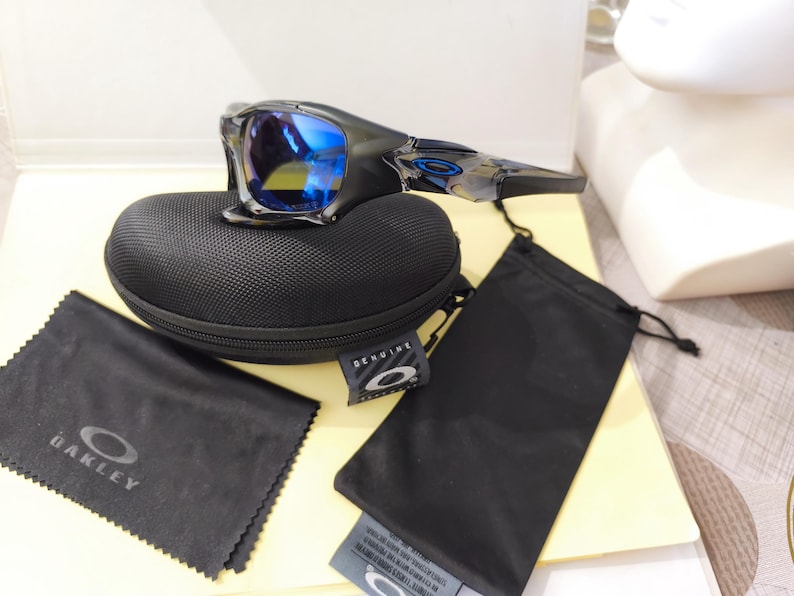 May include: Black Oakley sunglasses with blue mirrored lenses rest on a black case. A black microfiber cleaning cloth and a black drawstring bag are also present. The sunglasses and accessories are displayed on a light-colored surface.