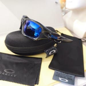 May include: Black and silver sunglasses with blue reflective lenses rest on a black case. Included are a black microfiber cleaning cloth with the Oakley logo and a black drawstring bag. A tag with the Oakley logo is also present.