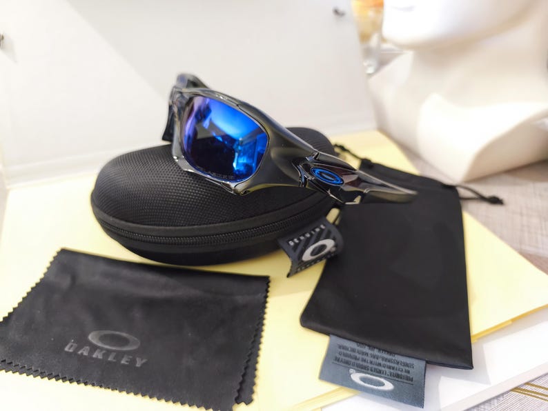 May include: Black and silver sunglasses with blue reflective lenses rest on a black case. Included are a black microfiber cleaning cloth with the Oakley logo and a black drawstring bag. A tag with the Oakley logo is also present.