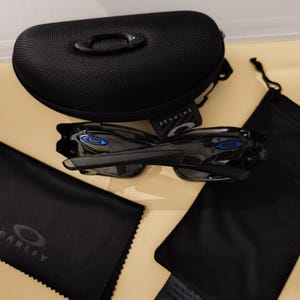 May include: Black Oakley sunglasses with blue accents, a black case, and several black pouches. The case has a textured surface and the word "GENUINE" is visible. The sunglasses are displayed with their accessories.