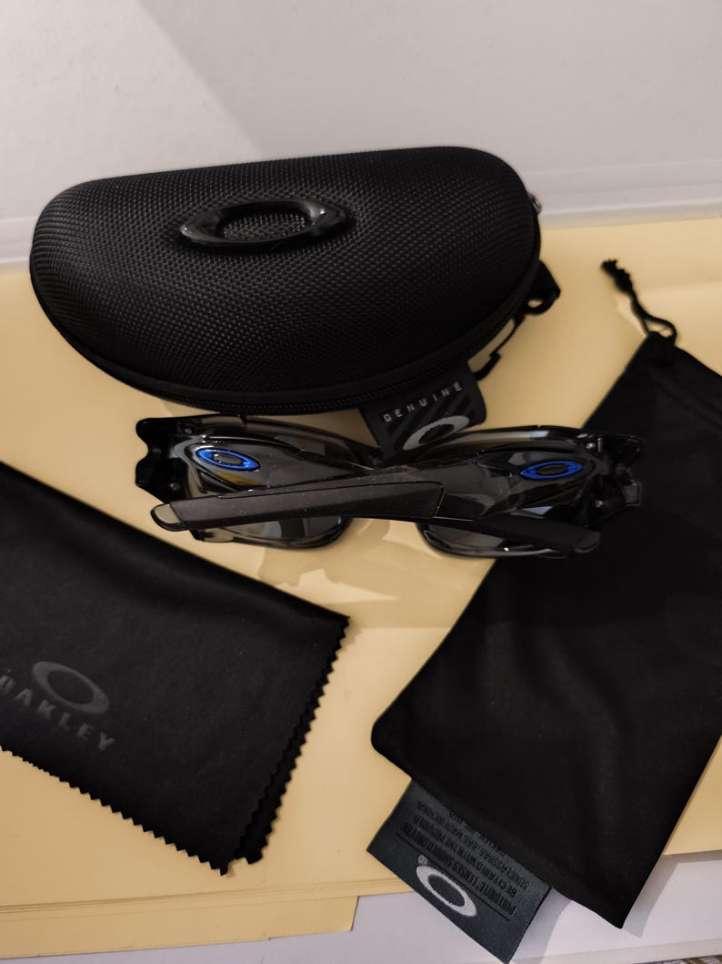 May include: Black Oakley sunglasses with blue accents, a black case, and several black pouches. The case has a textured surface and the word "GENUINE" is visible. The sunglasses are displayed with their accessories.