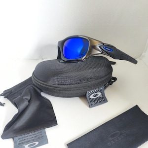May include: Black and silver sunglasses with blue lenses, resting on a black case. A black microfiber cleaning cloth and a black drawstring bag are also present. The sunglasses have a sleek, modern design.