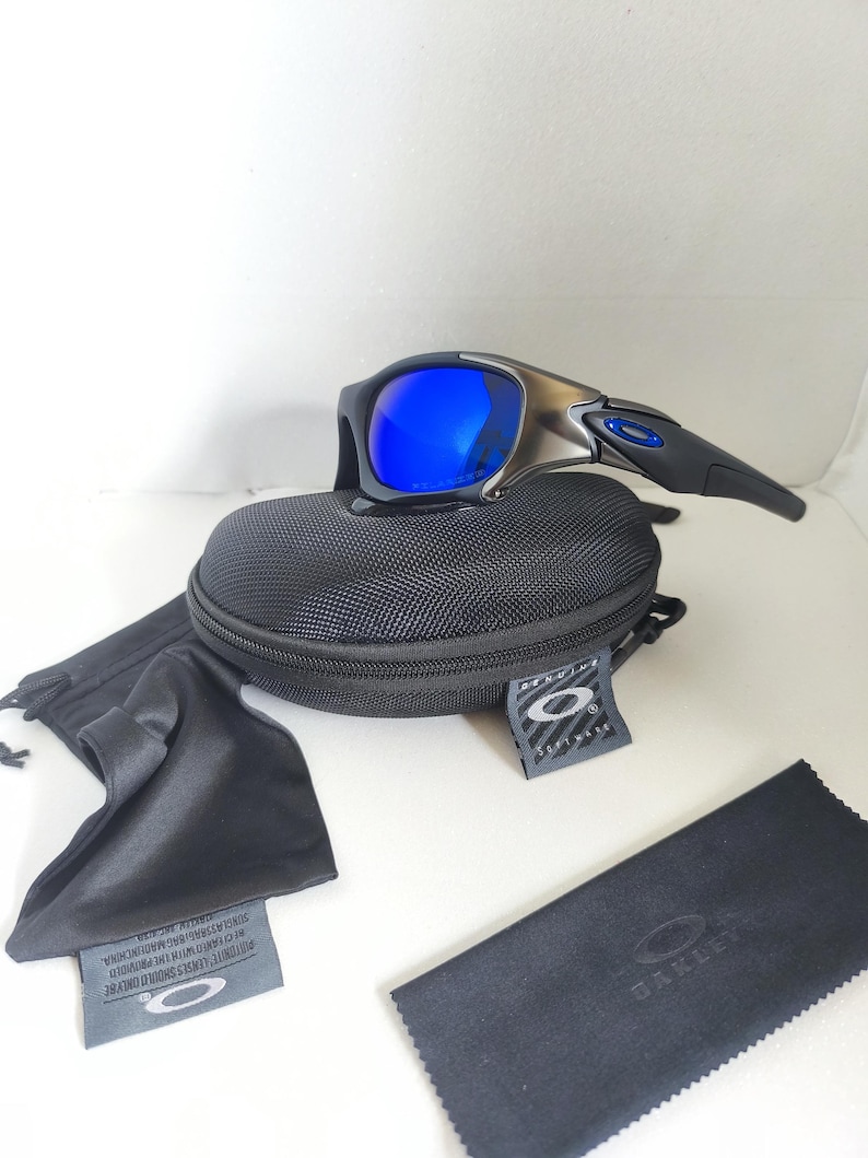 May include: Black and silver sunglasses with blue lenses, resting on a black case. A black microfiber cleaning cloth and a black drawstring bag are also present. The sunglasses have a sleek, modern design.