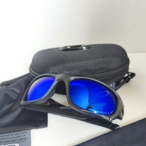 May include: Black sunglasses with blue mirrored lenses, a black case, and a cleaning cloth. The sunglasses have a sporty design and are likely for outdoor activities. The brand name "OAKLEY" is visible on the cloth.