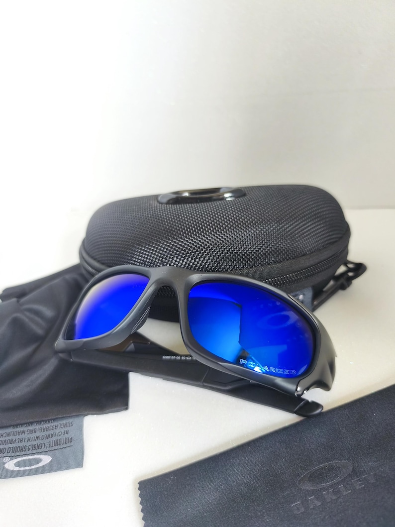 May include: Black sunglasses with blue mirrored lenses, a black case, and a cleaning cloth. The sunglasses have a sporty design and are likely for outdoor activities. The brand name "OAKLEY" is visible on the cloth.