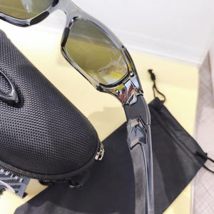 May include: Close-up of a pair of black and silver sunglasses with yellow-tinted lenses. The sunglasses are partially visible, along with a black carrying case and a black drawstring bag. The sunglasses have a modern, sporty design.