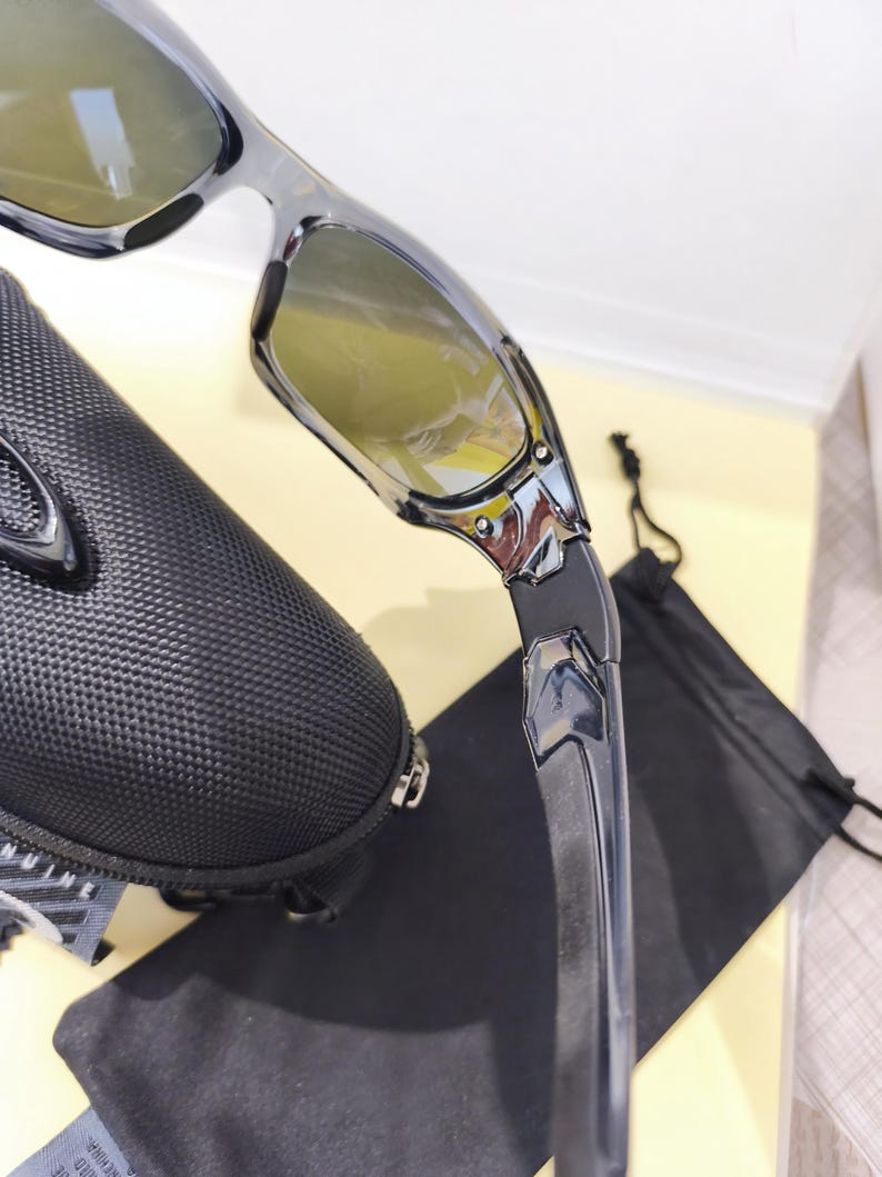 May include: Close-up of a pair of black and silver sunglasses with yellow-tinted lenses. The sunglasses are partially visible, along with a black carrying case and a black drawstring bag. The sunglasses have a modern, sporty design.
