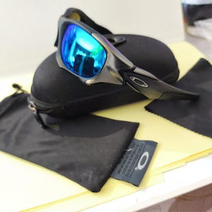 Puede incluir: Gafas de sol deportivas con lentes azules espejadas, sobre un estuche negro. La imagen incluye un pa&ntilde;o de microfibra negro y una peque&ntilde;a bolsa negra. Las gafas tienen una montura met&aacute;lica y un dise&ntilde;o elegante y moderno.