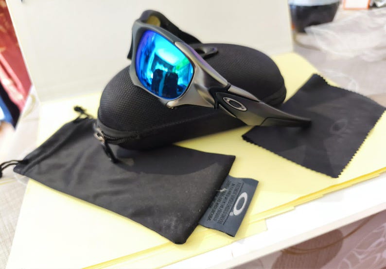 Puede incluir: Gafas de sol deportivas con lentes azules espejadas, sobre un estuche negro. La imagen incluye un pa&ntilde;o de microfibra negro y una peque&ntilde;a bolsa negra. Las gafas tienen una montura met&aacute;lica y un dise&ntilde;o elegante y moderno.
