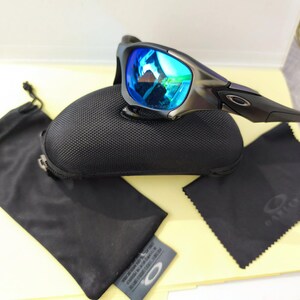 Puede incluir: Gafas de sol Oakley con lentes azules espejadas, sobre un estuche negro texturizado. Tambi&eacute;n se incluyen una bolsa negra con cord&oacute;n, un pa&ntilde;o de limpieza y una peque&ntilde;a etiqueta. Las gafas tienen una montura gris oscuro.