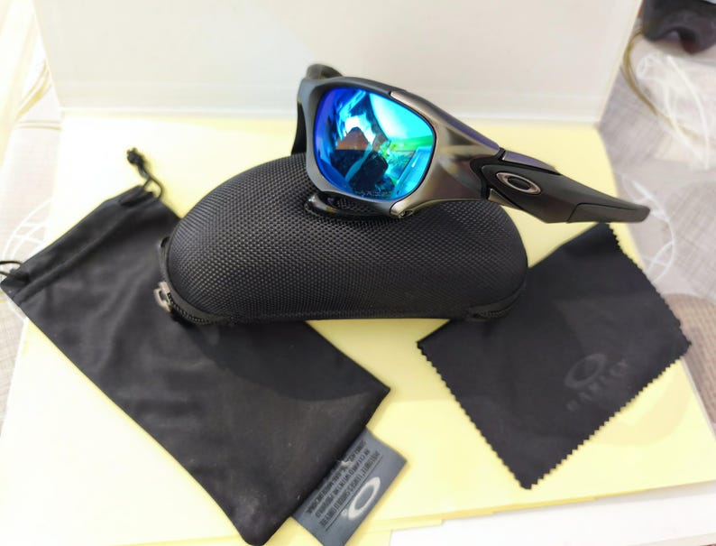 Puede incluir: Gafas de sol Oakley con lentes azules espejadas, sobre un estuche negro texturizado. Tambi&eacute;n se incluyen una bolsa negra con cord&oacute;n, un pa&ntilde;o de limpieza y una peque&ntilde;a etiqueta. Las gafas tienen una montura gris oscuro.