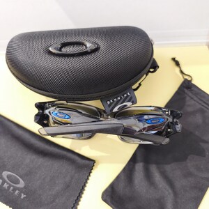 May include: Black Oakley sunglasses with blue accents, a black case, and cleaning cloths. The sunglasses have a sleek, modern design. The case is textured and features the Oakley logo. The cleaning cloths are black.