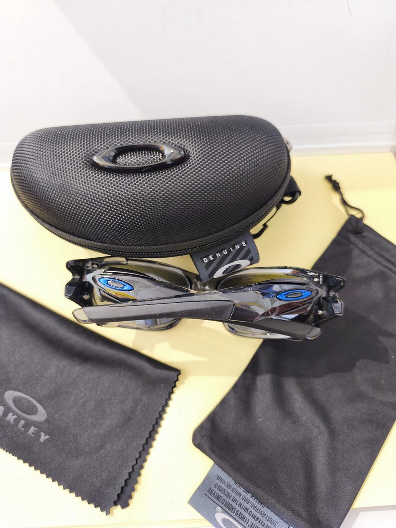 May include: Black Oakley sunglasses with blue accents, a black case, and cleaning cloths. The sunglasses have a sleek, modern design. The case is textured and features the Oakley logo. The cleaning cloths are black.