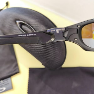 Puede incluir: Gafas de sol negras Oakley Pit Boss II con lentes de color naranja. Las gafas de sol tienen un acabado mate y el texto "PIT BOSS II" es visible en la montura. La imagen tambi&eacute;n muestra un estuche negro, un pa&ntilde;o de limpieza y una peque&ntilde;a etiqueta.