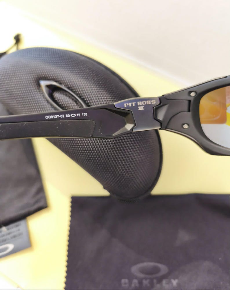 Puede incluir: Gafas de sol negras Oakley Pit Boss II con lentes de color naranja. Las gafas de sol tienen un acabado mate y el texto "PIT BOSS II" es visible en la montura. La imagen tambi&eacute;n muestra un estuche negro, un pa&ntilde;o de limpieza y una peque&ntilde;a etiqueta.