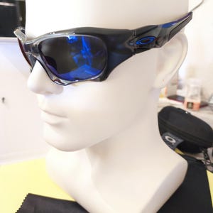 May include: Black and blue sunglasses with a glossy finish are displayed on a white mannequin head. The lenses reflect a blue hue. The sunglasses have a sporty design, with the brand logo visible on the temples.