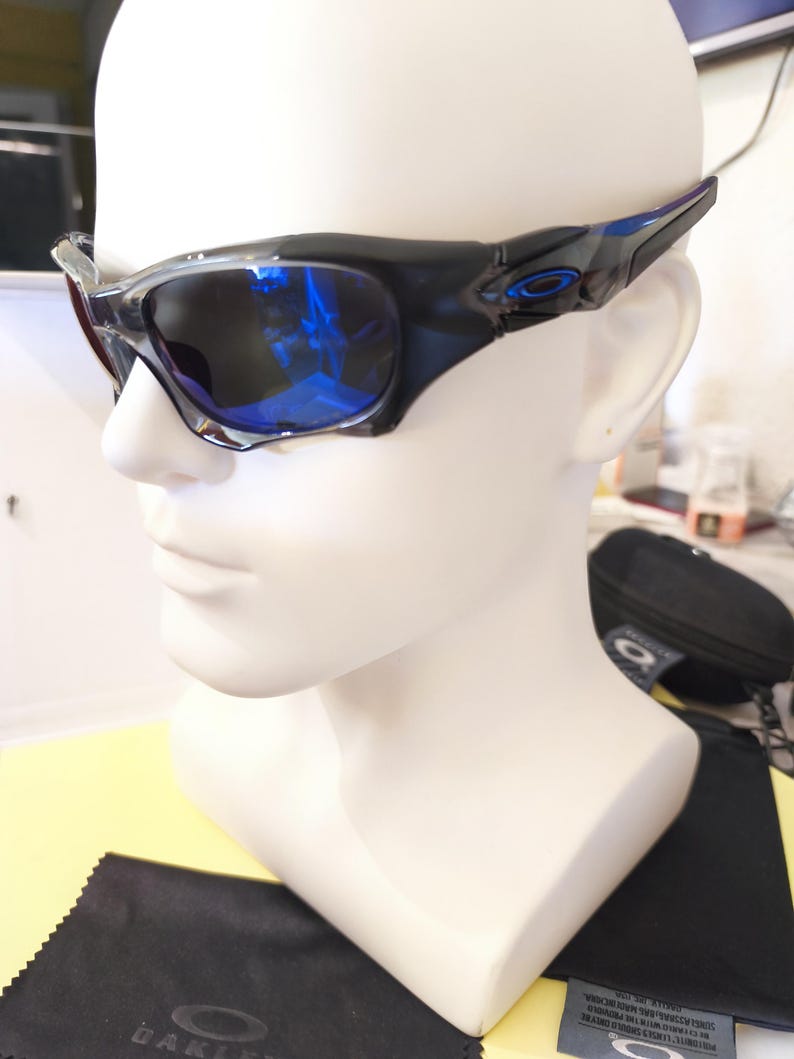May include: Black and blue sunglasses with a glossy finish are displayed on a white mannequin head. The lenses reflect a blue hue. The sunglasses have a sporty design, with the brand logo visible on the temples.