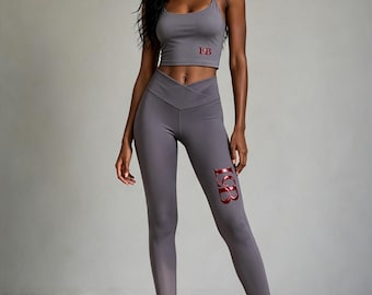 May include: A grey two-piece athletic set consisting of a cropped tank top with thin straps and high-waisted leggings. Both pieces feature the red metallic letters LGB. The leggings have a V-shaped crossover waistband and a fitted silhouette.