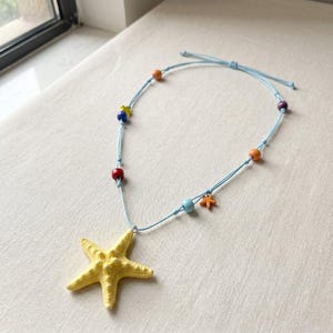 May include: A light blue cord necklace featuring a large yellow starfish pendant. The cord is accented with small, colorful beads in shades of orange, blue, red, and purple, along with a tiny orange starfish charm attached to the side of the cord.