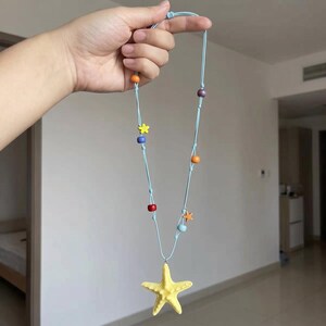 May include: A light blue cord necklace featuring a large yellow starfish pendant at the center. The cord is decorated with several small, colorful beads in shades of orange, red, purple, and light blue, along with two tiny yellow and orange starfish charms.