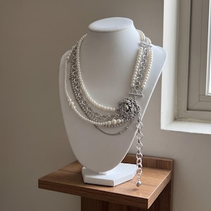 May include: A multi-strand layered necklace featuring white pearls, silver-toned metal chains, and a row of clear rhinestones. The strands are connected by a circular silver-toned ornate medallion and a long dangling chain with a spherical charm.