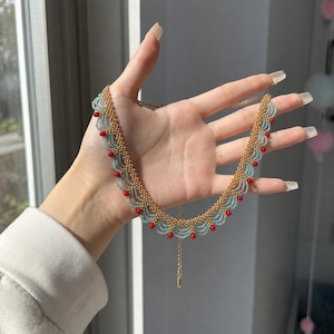 May include: A gold-coloured beaded necklace with a scalloped design. The necklace has light blue and red beads. The necklace is held up in front of a window, showing the intricate details of the design.