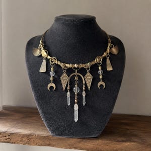 Op de afbeelding: Een goudkleurige statement ketting met hemelse en geometrische bedels. De ketting heeft halve manen, kristallen hangers en ruitvormige bedels met oogontwerpen. De ketting wordt getoond op een zwarte mannequin.