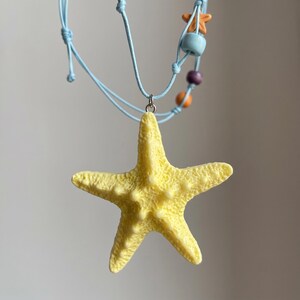 May include: A light blue cord necklace featuring a large, textured yellow starfish pendant. The cord is accented with small, colorful beads in shades of orange, light blue, and purple, suspended from a silver-toned metal jump ring.
