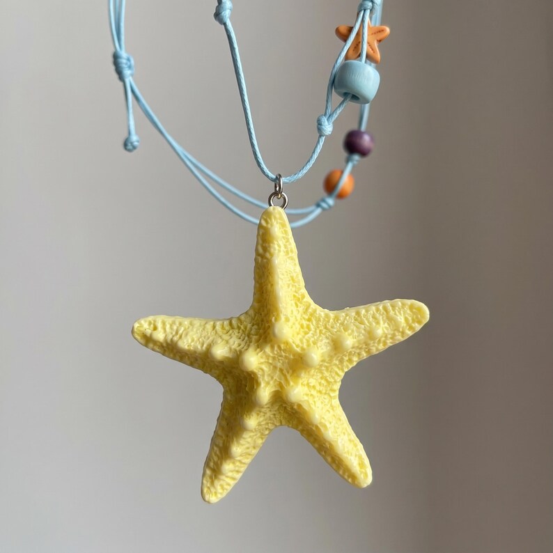 May include: A light blue cord necklace featuring a large, textured yellow starfish pendant. The cord is accented with small, colorful beads in shades of orange, light blue, and purple, suspended from a silver-toned metal jump ring.