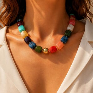 May include: A necklace featuring a series of multi-colored, square-shaped beads in shades of red, blue, green, yellow, and purple, interspersed with a single, central, polished gold-tone spherical bead, all strung together on a cord.