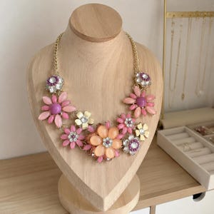 Può includere: Una collana importante con una catena color oro decorata da un gruppo di ciondoli a forma di fiore. I fiori sono realizzati con petali rosa, color pesca e crema con accenti centrali di perline viola e strass trasparenti scintillanti.