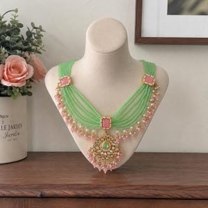 Op de afbeelding: Een mintgroene kralenketting met roze accenten en een gouden hanger. De ketting wordt op een mannequin getoond. De hanger heeft een centrale groene steen en is omgeven door roze kralen en gouden details. De ketting is een statement piece.
