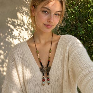 Peut inclure: Un long collier avec un pendentif papillon central de couleur bronze. Le cordon est orné de diverses perles, notamment des sphères noires lisses, des sphères rouges, des sphères vert clair et des perles cylindriques couleur os.