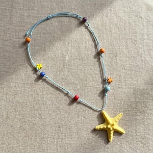 May include: A light blue cord necklace featuring a large yellow starfish pendant. The necklace is accented with small, colorful beads in shades of purple, orange, blue, red, and yellow, along with a small yellow flower charm and a tiny orange starfish.