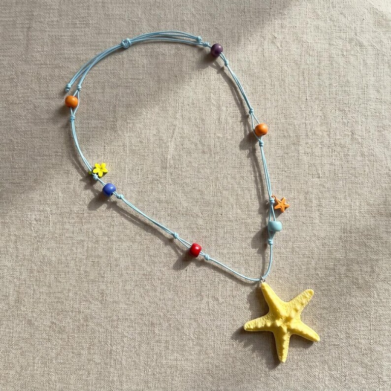 May include: A light blue cord necklace featuring a large yellow starfish pendant. The necklace is accented with small, colorful beads in shades of purple, orange, blue, red, and yellow, along with a small yellow flower charm and a tiny orange starfish.