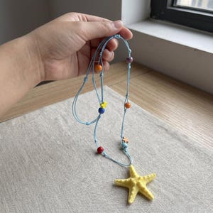 May include: A light blue cord necklace featuring a large yellow starfish pendant. The cord is accented with small, colorful beads in shades of red, orange, blue, purple, and yellow, along with tiny star-shaped charms spaced along the length.