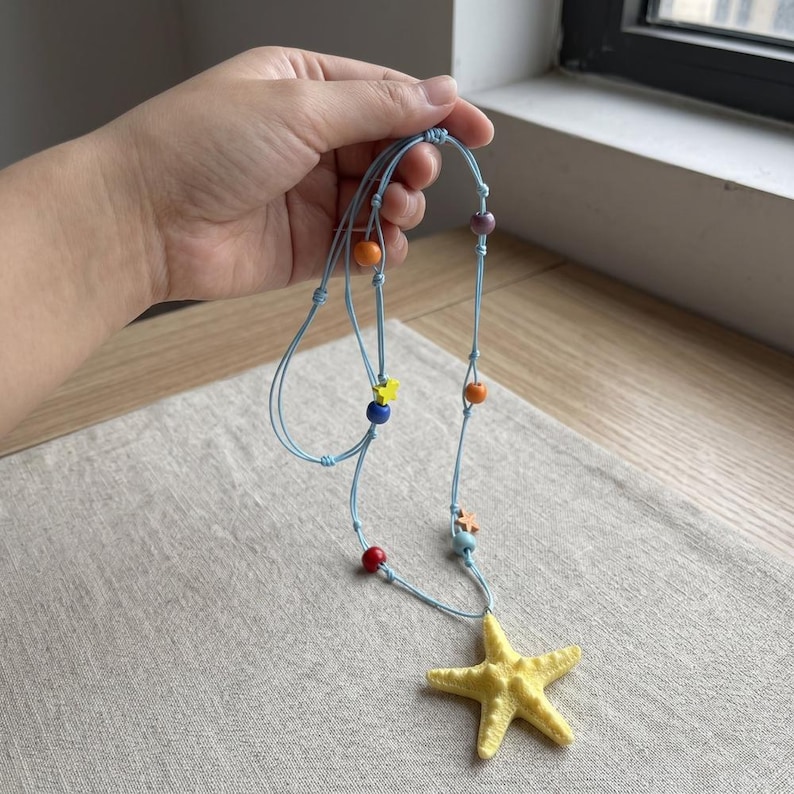 May include: A light blue cord necklace featuring a large yellow starfish pendant. The cord is accented with small, colorful beads in shades of red, orange, blue, purple, and yellow, along with tiny star-shaped charms spaced along the length.