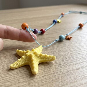 May include: A necklace featuring a large, textured yellow starfish pendant suspended from a light blue cord. The cord is accented with several small, round, multi-colored beads in shades of orange, purple, red, blue, and yellow, along with a tiny star charm.