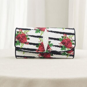 May include: A rectangular clutch bag featuring a textured white surface with horizontal black stripes and a vibrant red rose floral pattern. The center of the flap displays a distinct, elongated, diamond-shaped white textured accent piece.