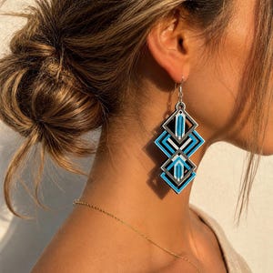 Puede incluir: Pendientes colgantes geométricos con un gancho plateado. Los pendientes presentan un diseño en capas de formas geométricas negras, grises y azul turquesa. Un accesorio moderno.