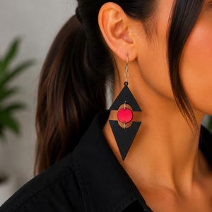 Puede incluir: Un pendiente colgante geométrico que presenta dos formas triangulares de color negro mate conectadas por un disco circular rojo brillante en el centro. El pendiente cuelga de un gancho de metal plateado, creando un diseño angular y superpuesto.