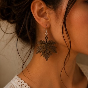 Peut inclure: Une boucle d'oreille en forme de feuille d'érable brun foncé avec une texture de grain de bois détaillée et un crochet en métal argenté. La forme de la feuille est complexe avec des bords pointus et une tige centrale, suspendue à une boucle.