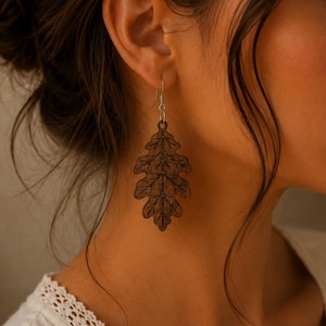 May include: A dark brown wooden earring shaped like an oak leaf with detailed laser-engraved vein patterns. The leaf pendant hangs from a simple silver-toned metal hook wire, creating a natural and organic appearance for this dangling jewelry piece.