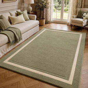 May include: A rectangular area rug featuring a solid sage green center field framed by a wide, cream-colored border. The rug has a textured, low-pile surface with a clean, straight-edged finish, providing a neutral foundation for interior spaces.