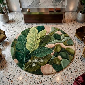Puede incluir: Una alfombra circular con un dise&ntilde;o de mechones de grandes hojas tropicales superpuestas en tonos verde oscuro y claro, intercaladas con formas redondeadas y suaves similares a guijarros en tonos verde y beige. El texto finehandicrafts gifts es visible.