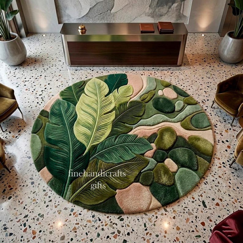 Puede incluir: Una alfombra circular con un dise&ntilde;o de mechones de grandes hojas tropicales superpuestas en tonos verde oscuro y claro, intercaladas con formas redondeadas y suaves similares a guijarros en tonos verde y beige. El texto finehandicrafts gifts es visible.
