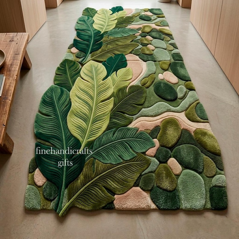 Puede incluir: Una alfombra rectangular con un dise&ntilde;o texturizado de grandes hojas tropicales superpuestas en varios tonos de verde y beige claro, intercaladas con formas redondeadas similares a piedras. El texto finehandicrafts gifts est&aacute; impreso en la alfombra.